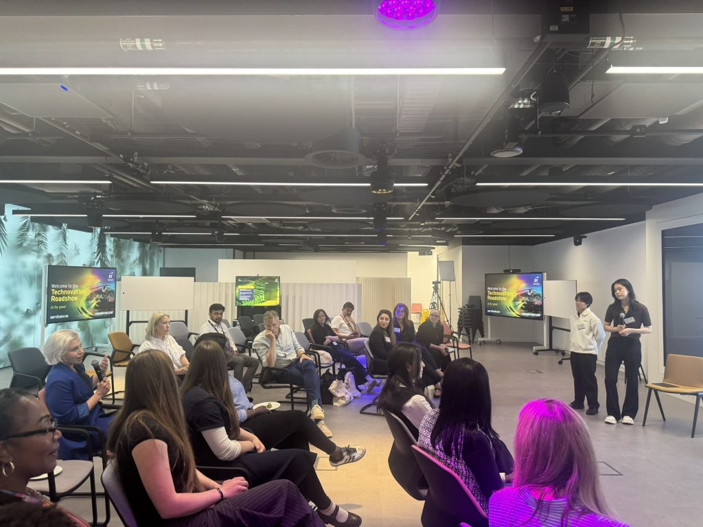 Girls and other attendees sit in a circle as two participants stand to present their idea during an interactive session at the Technovation London Roadshow.